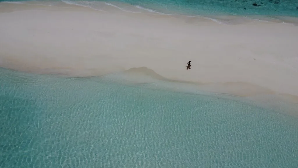 Luftaufnahme einer Person, die entlang einer unberührten Sandbank auf den Malediven spaziert, umgeben von türkisblauem Wasser.