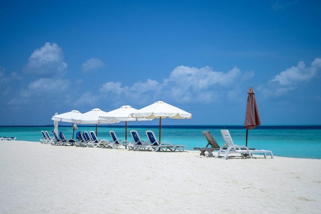 Bikini Beach in Gulhi, Malediven, mit weißem Sand, türkisfarbenem Wasser und Sonnenliegen unter Palmen.