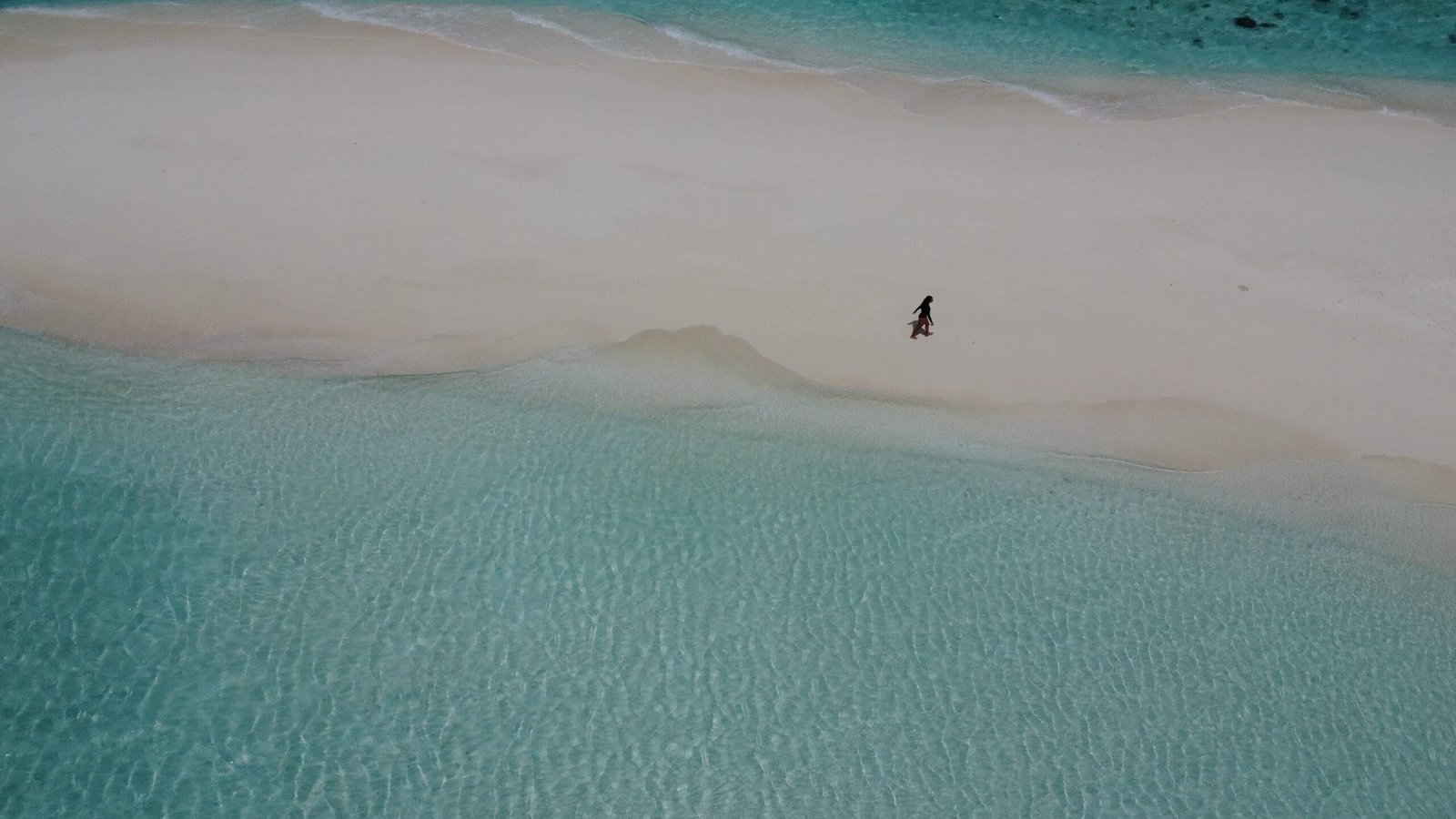 Aerial view of a person strolling along a pristine sandbank beach in the Maldives, surrounded by turquoise waters.